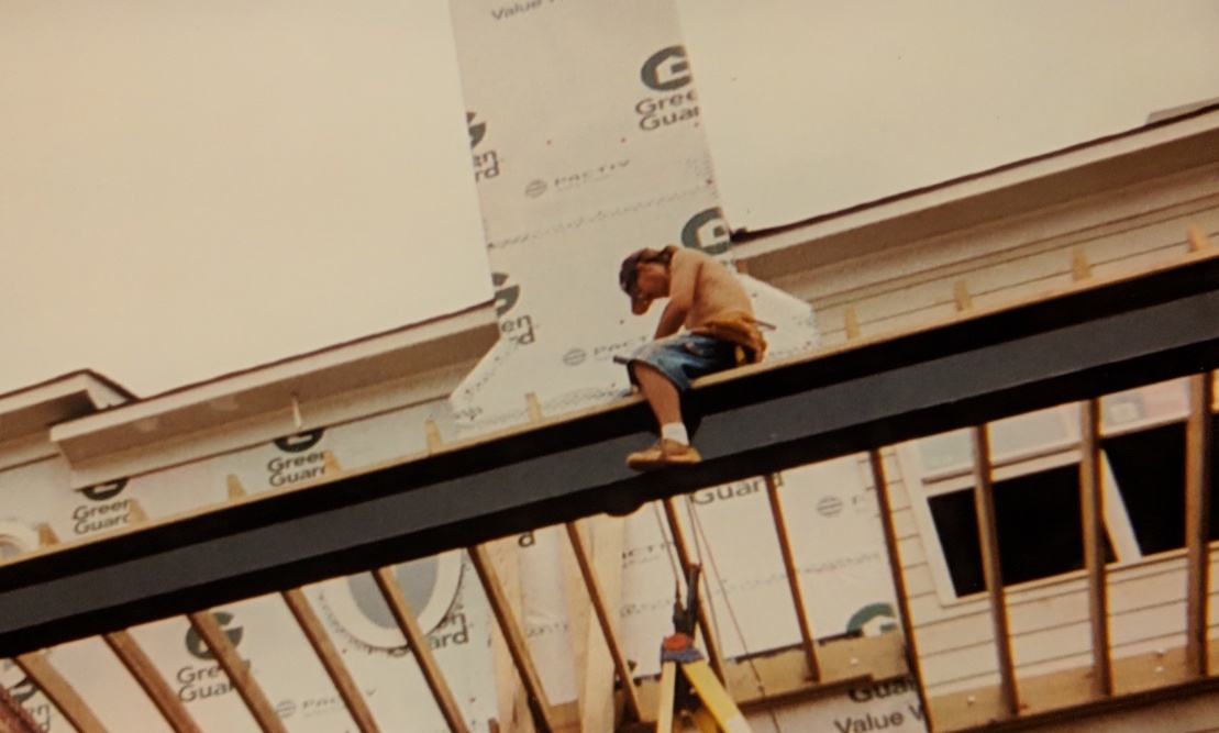 construction associate sitting atop one of the horizontal beams of a second story deck being built alongside a chimney